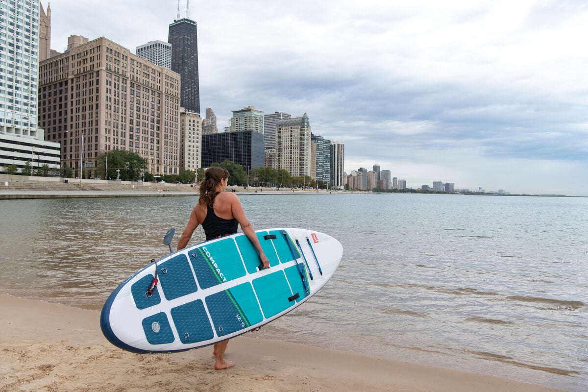 Woman carrying compact inflatable paddle board down the beach toward the water