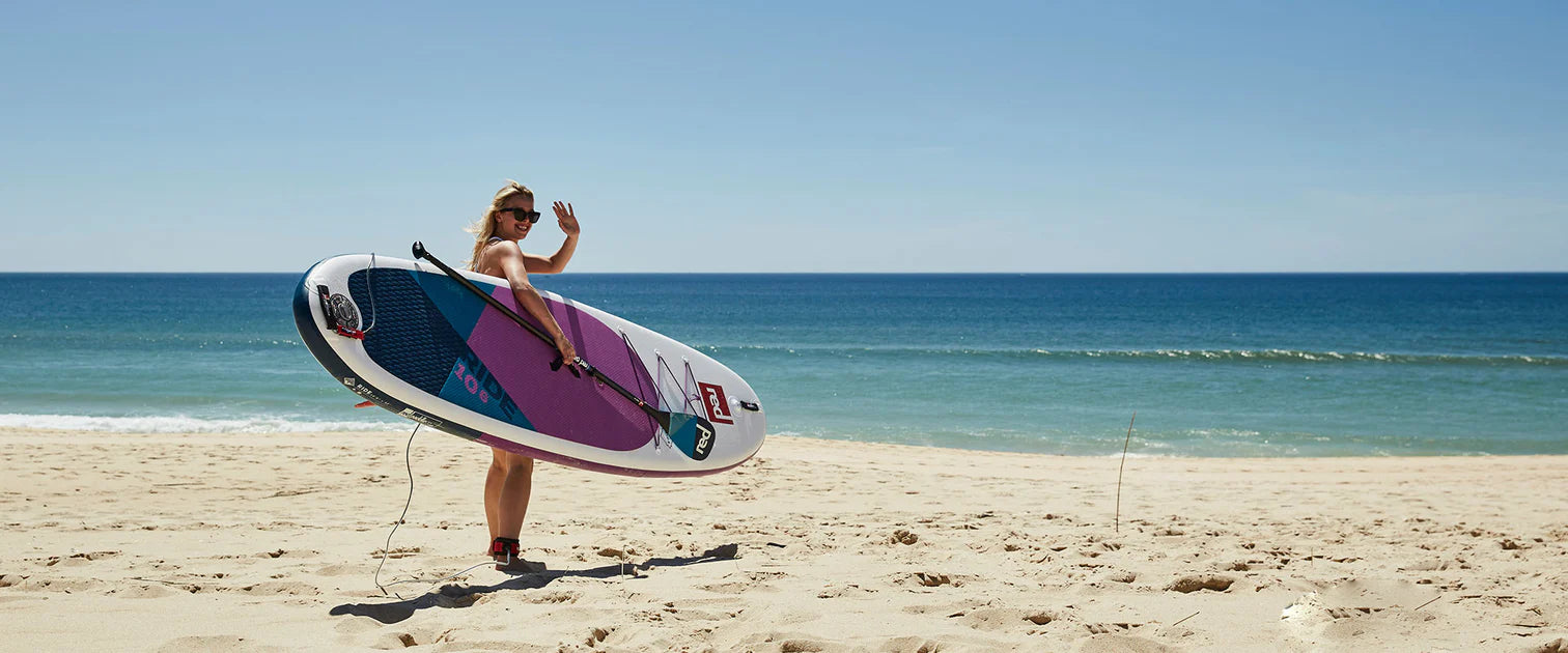 A woman on the beach, carrying her paddleboard, waving at someone