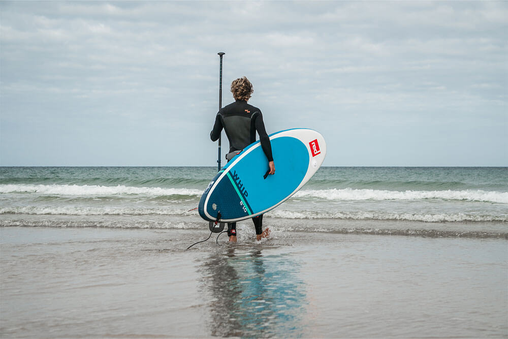 man wearing a wetsuit walking to the sea carrying Whip MSL inflatable paddle board