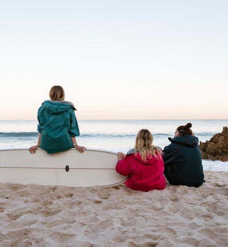 Three women sat on the beach next to a surf board wearing Red Equipment Pro Change Robe EVO