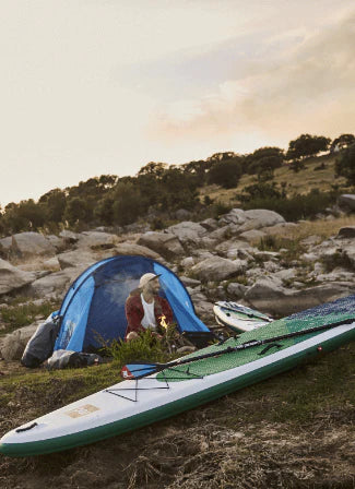 A man camping, sat in front of a fire with a paddleboard in the foreground